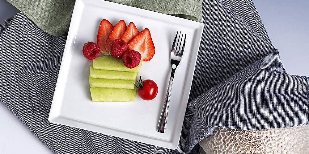 Cameo China Narrow Rim white square plate featuring elegant fruit presentation with sliced strawberries, raspberries, kiwi fan and cherry tomato with fork on striped linens